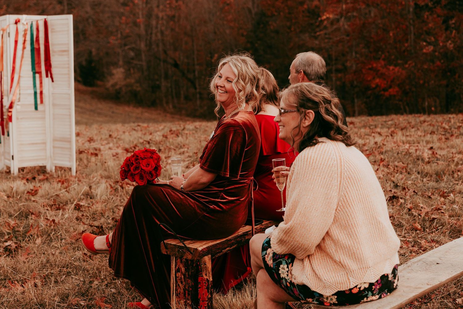 Smiling wedding guests in red and floral attire seated on wooden benches at an outdoor autumn wedding ceremony.