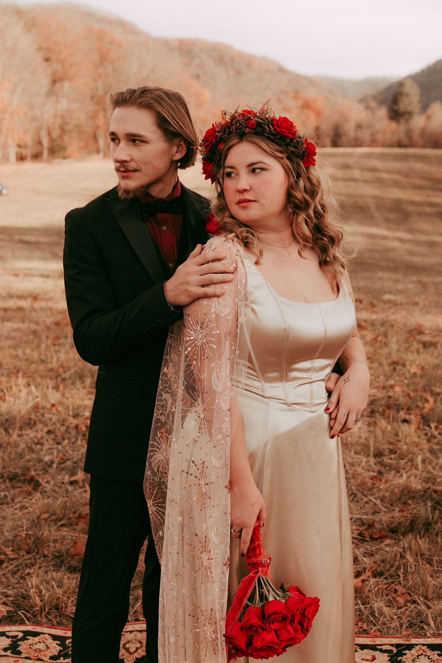 Wedding couple posing in mountain field, groom in black suit, bride in white gown with red floral crown.