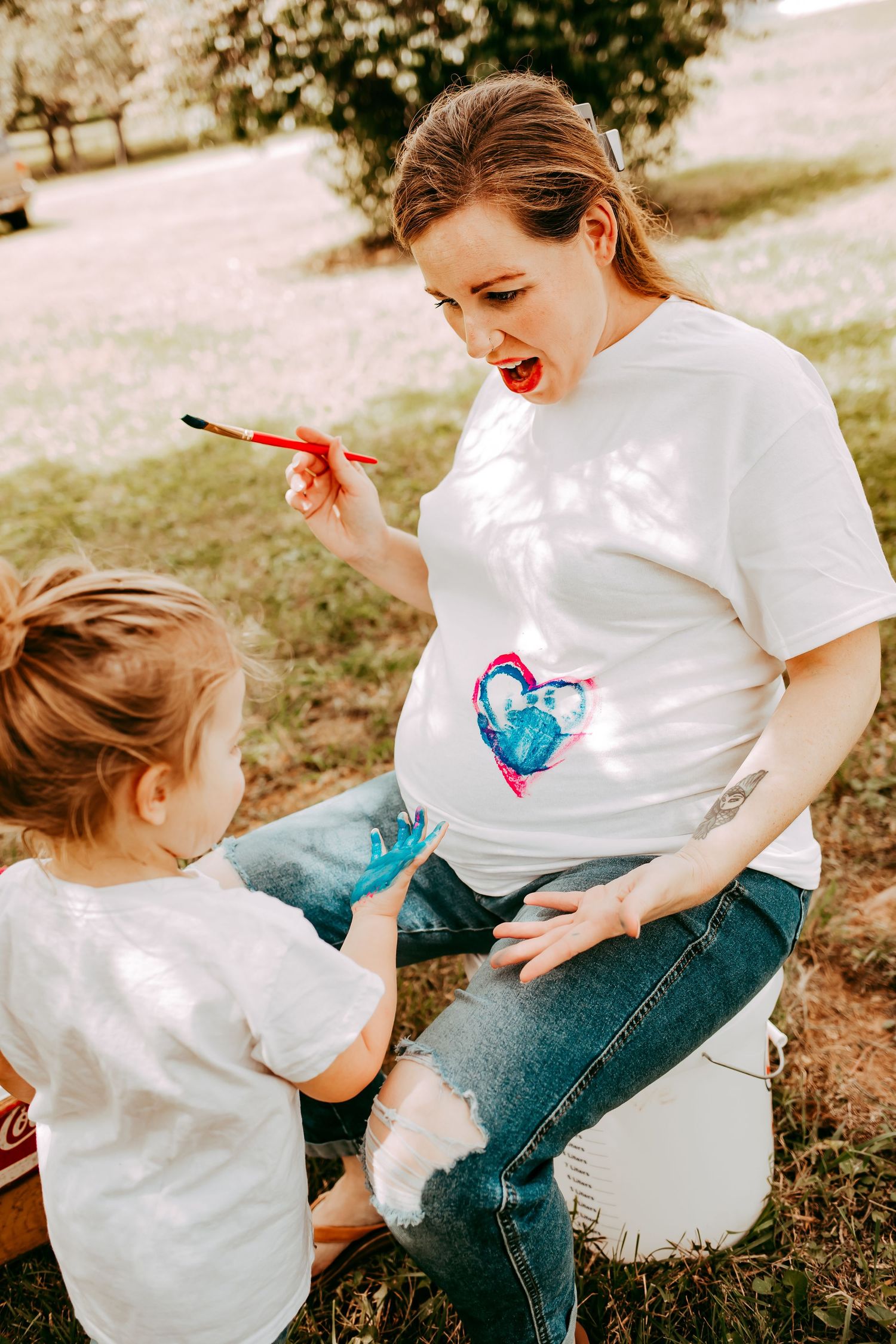 A laughing pregnant woman playfully holds a paintbrush while her young child touches her painted baby bump outside.