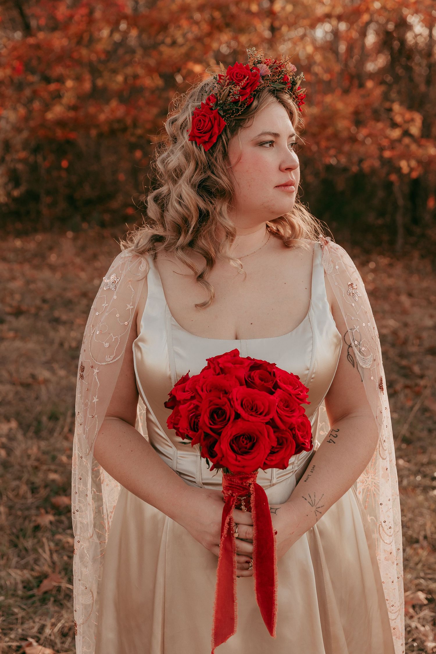 Bride holds red rose bouquet wearing gold gown and floral crown in fall setting.