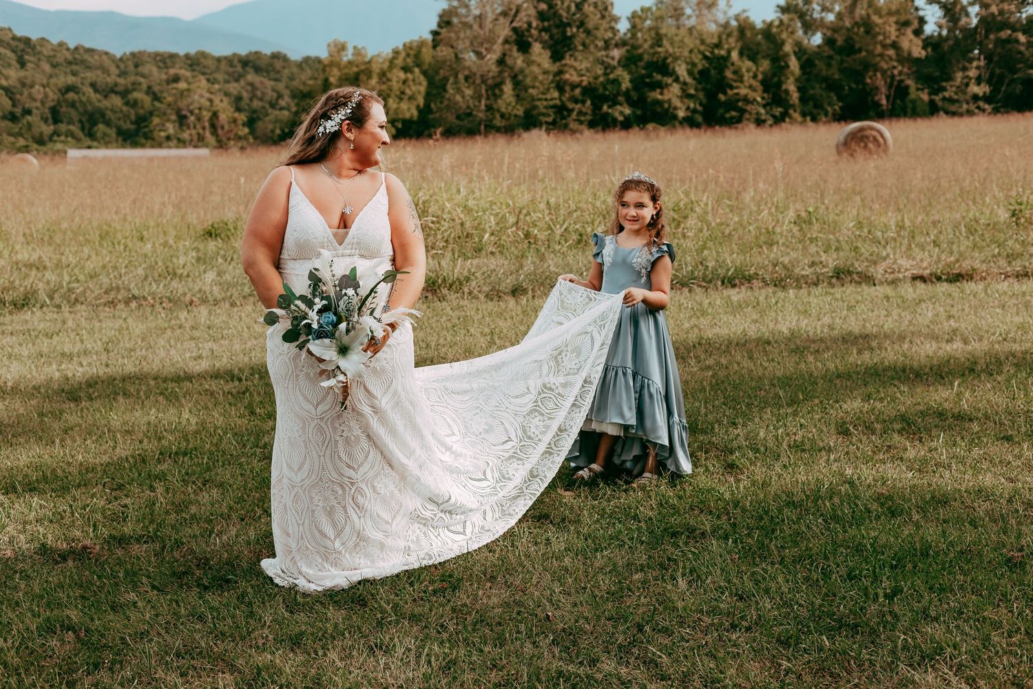 Bride in lace gown walks with young flower girl in blue dress across a sunlit meadow wedding venue.