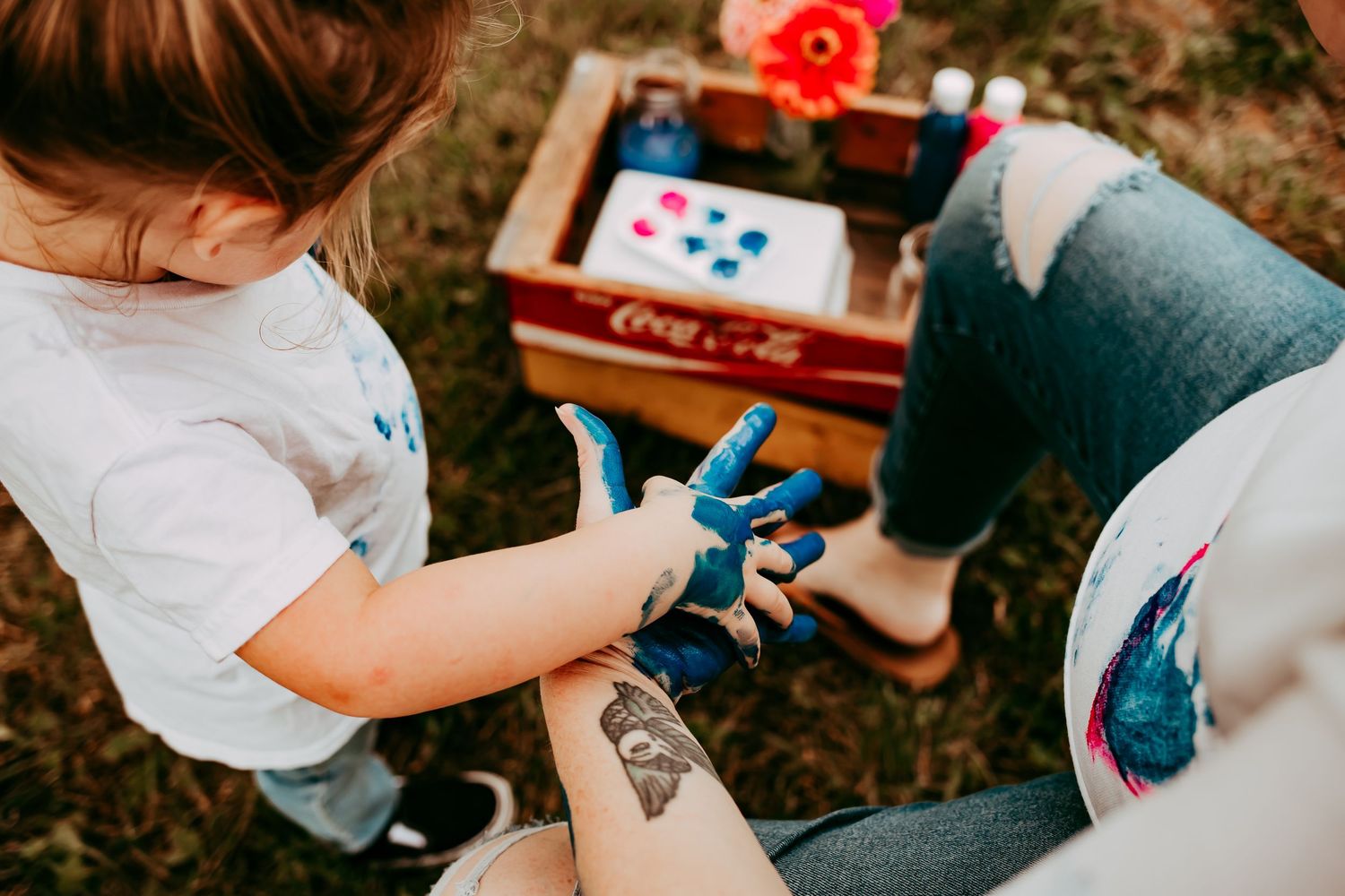 A toddler with blue paint-covered hands sits beside a pregnant woman with a henna tattoo outdoors.