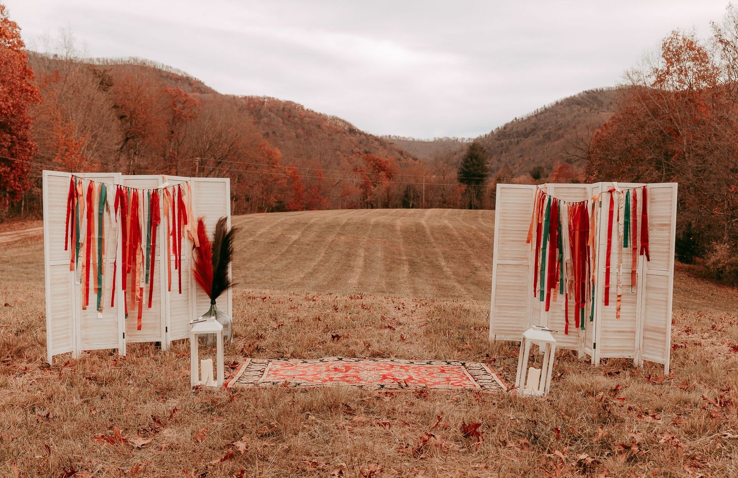 Rustic outdoor fall wedding ceremony backdrop with white shutters adorned with colorful ribbon streamers in mountain landscape.