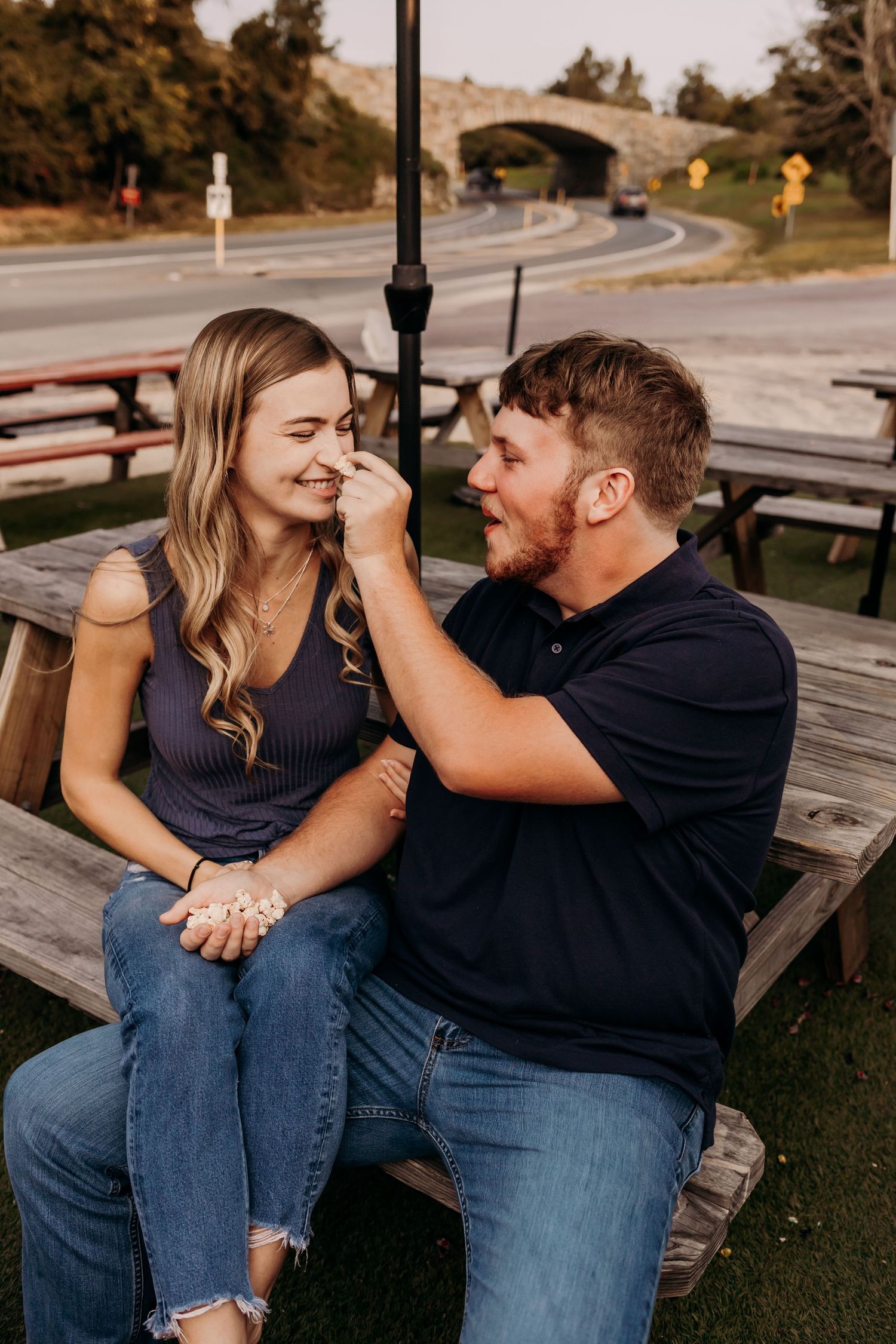 Couple sitting on picnic bench, man playfully touching woman's nose while she laughs outdoors.