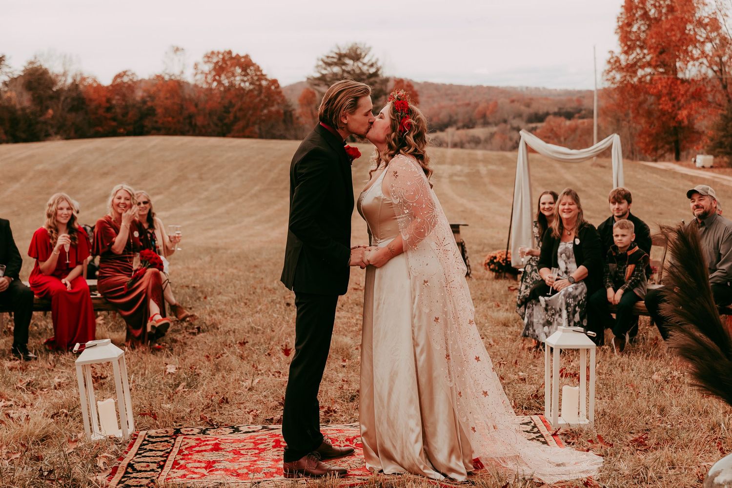 Bride and groom sharing first kiss at outdoor fall wedding ceremony with guests seated in field.