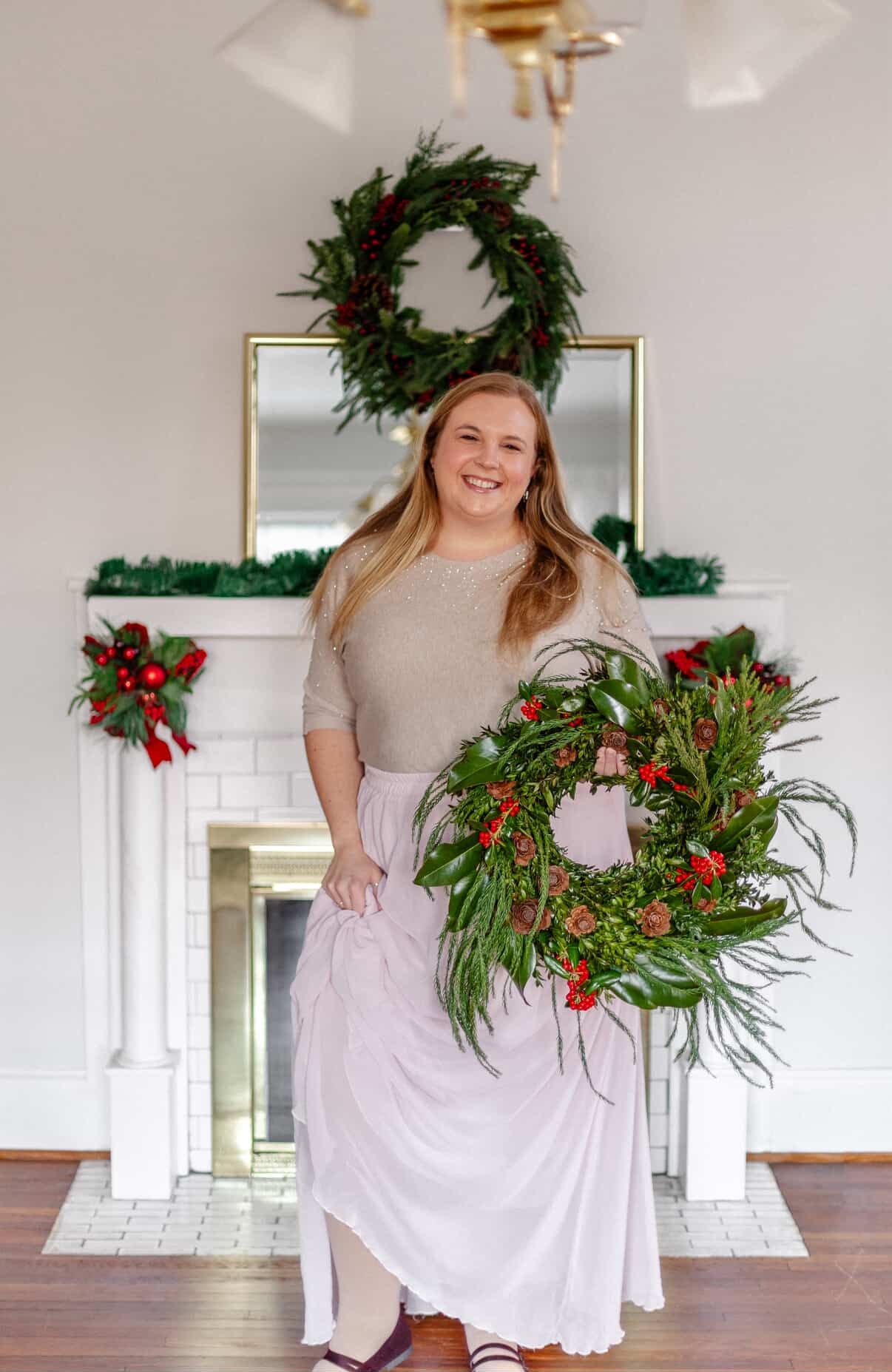 Smiling woman holding a festive Christmas wreath in front of a decorated holiday fireplace mantel.