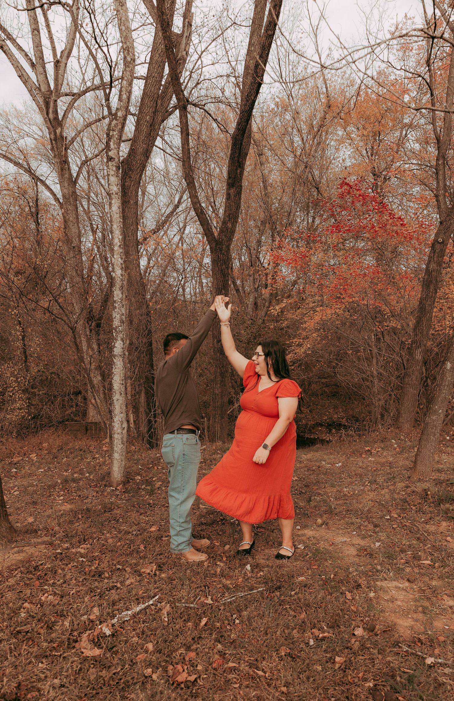 Couple dancing outdoors in autumn forest, woman in red dress twirling, surrounded by bare trees and fall foliage.