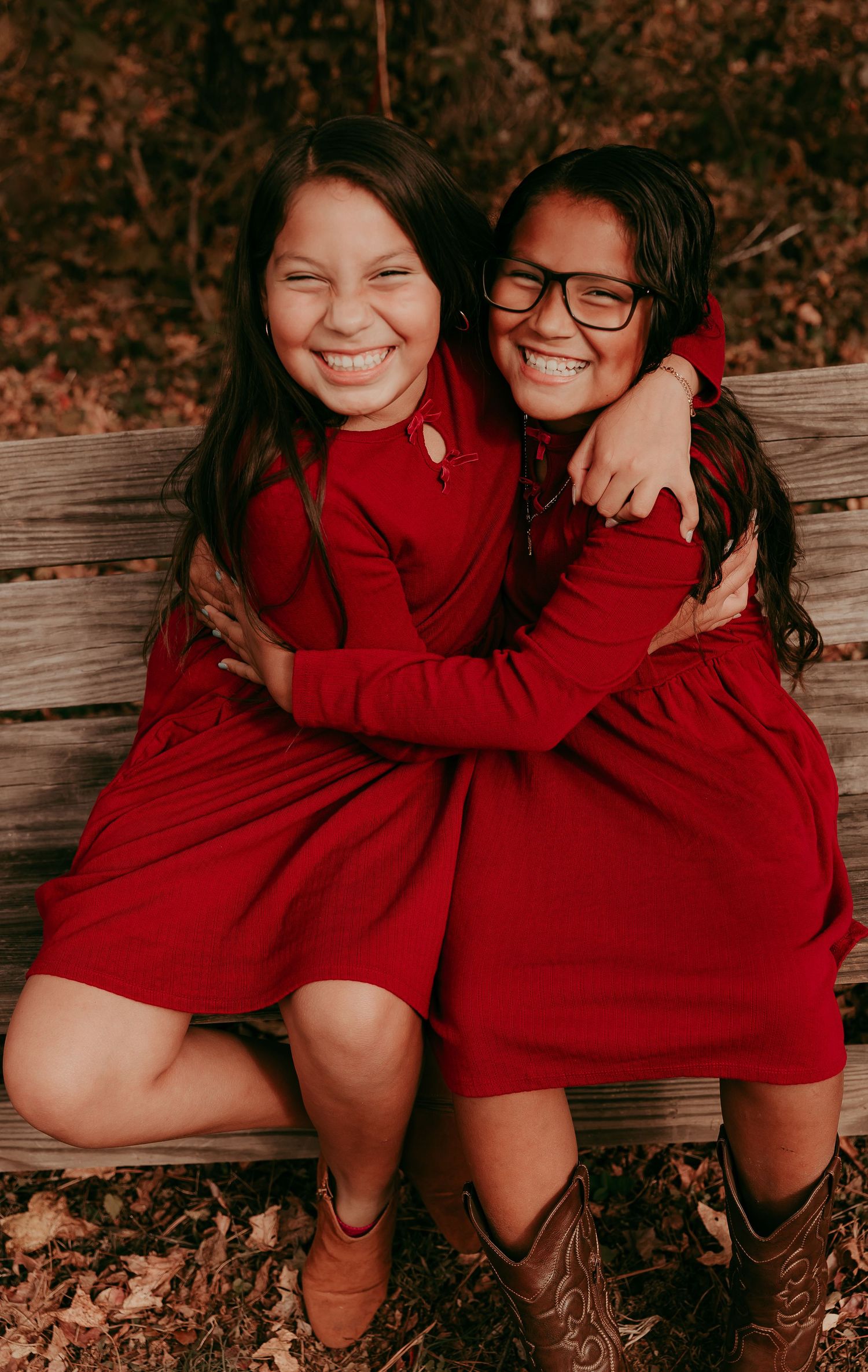 Two girls in matching red dresses hugging and smiling on a wooden bench.