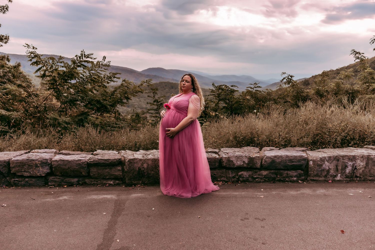 Pregnant woman in flowing pink tulle dress standing at scenic mountain overlook at dusk.