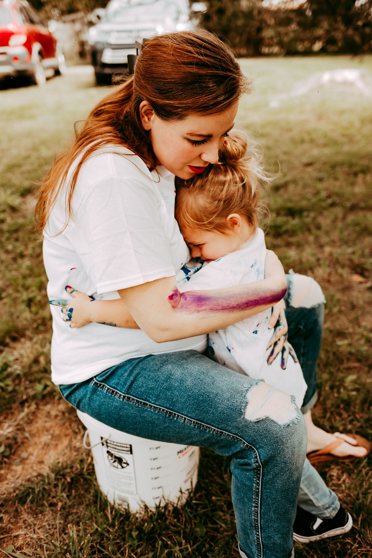 A pregnant mother tenderly hugs her sleepy toddler while sitting outdoors in a grassy field.