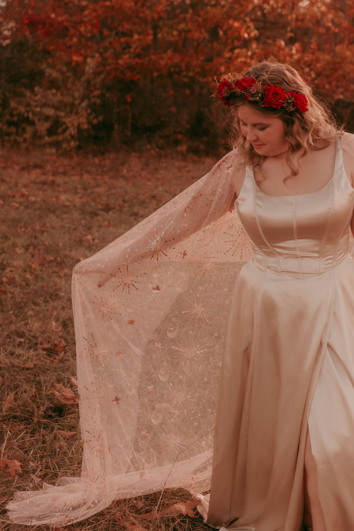 Bride in gold satin gown displays embroidered lace cape in autumn forest setting.