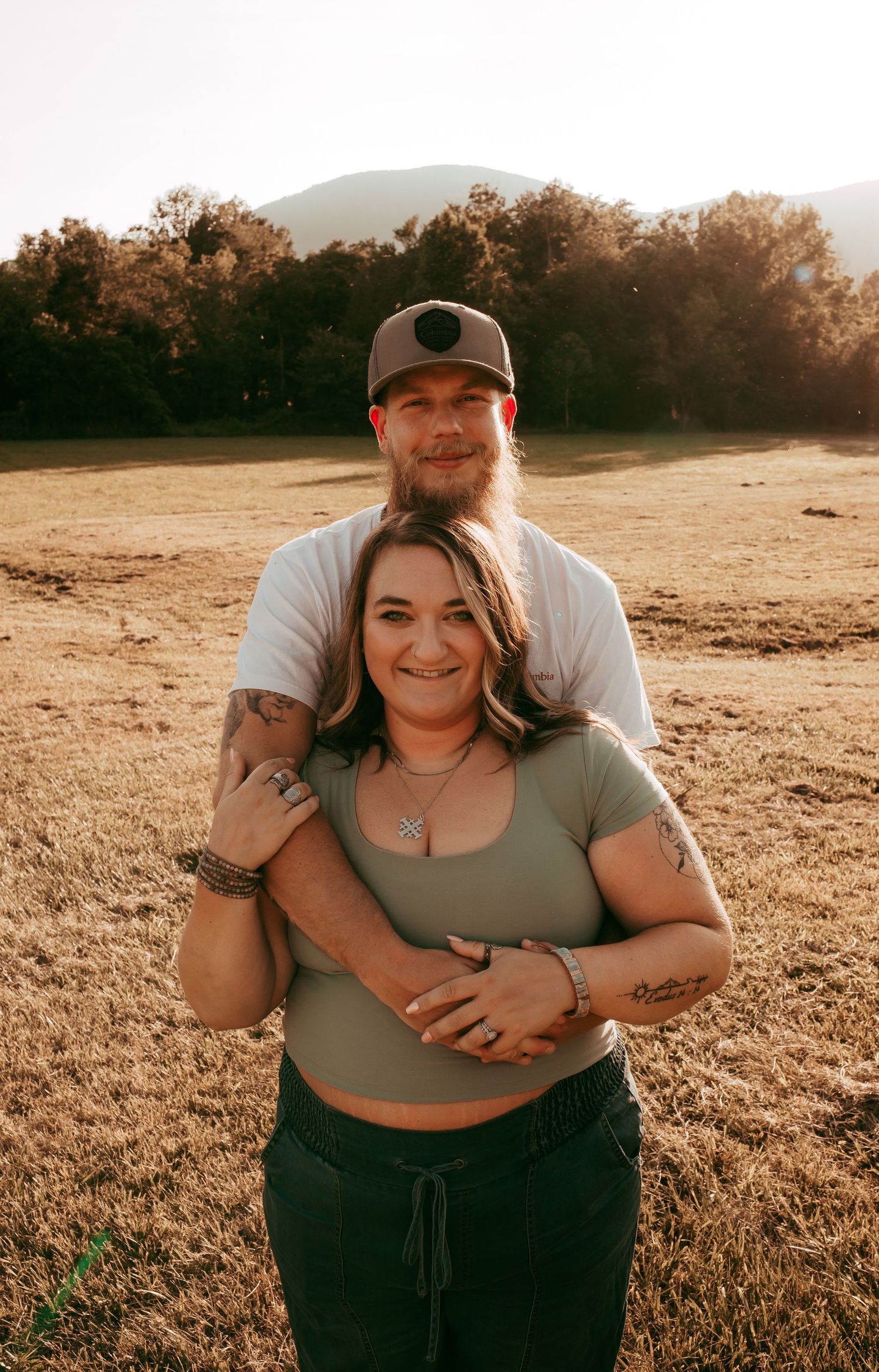 Smiling couple posing together in warm golden hour light in open field with mountain and tree backdrop.