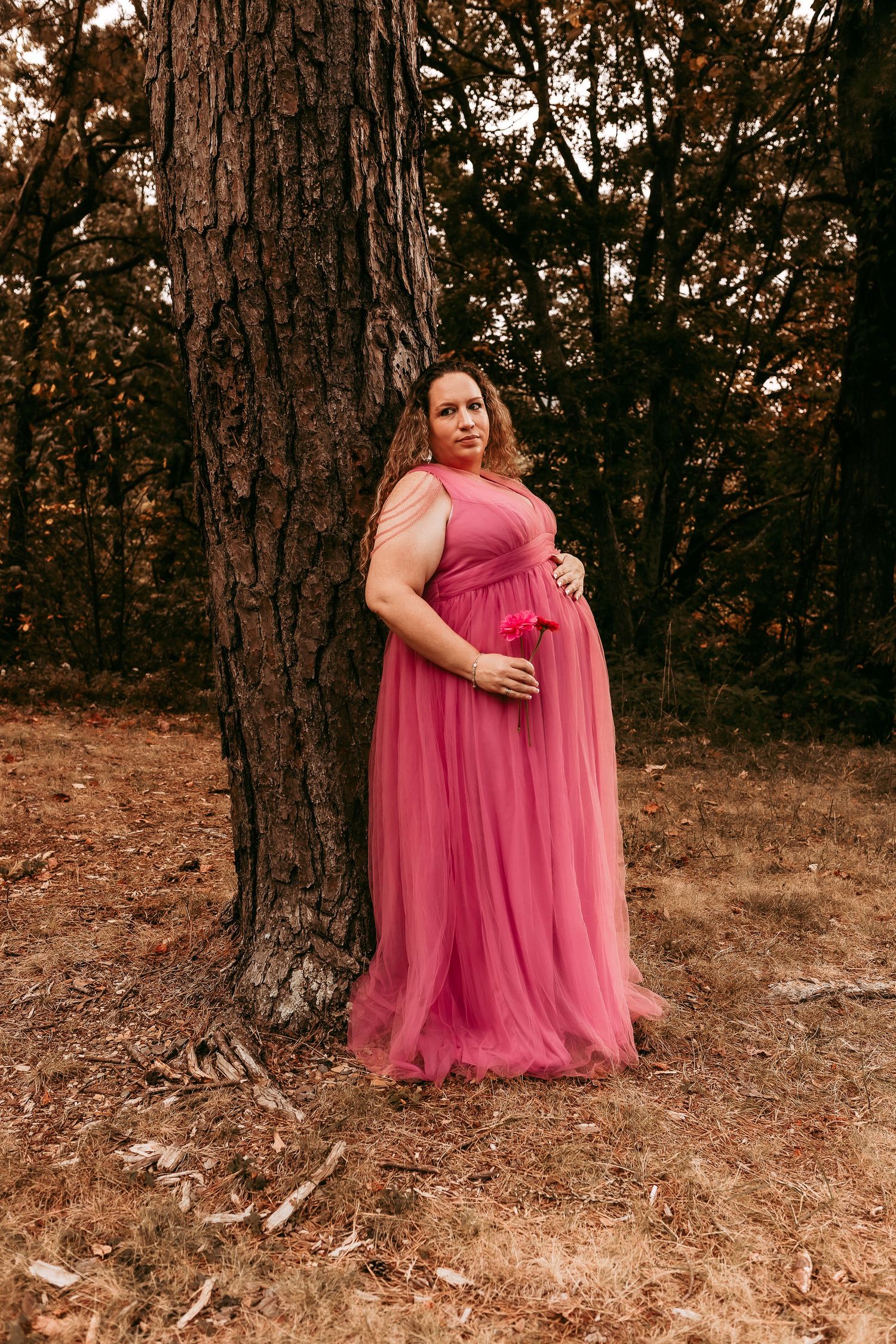Pregnant woman in pink tulle gown leaning against large tree trunk in autumn forest.