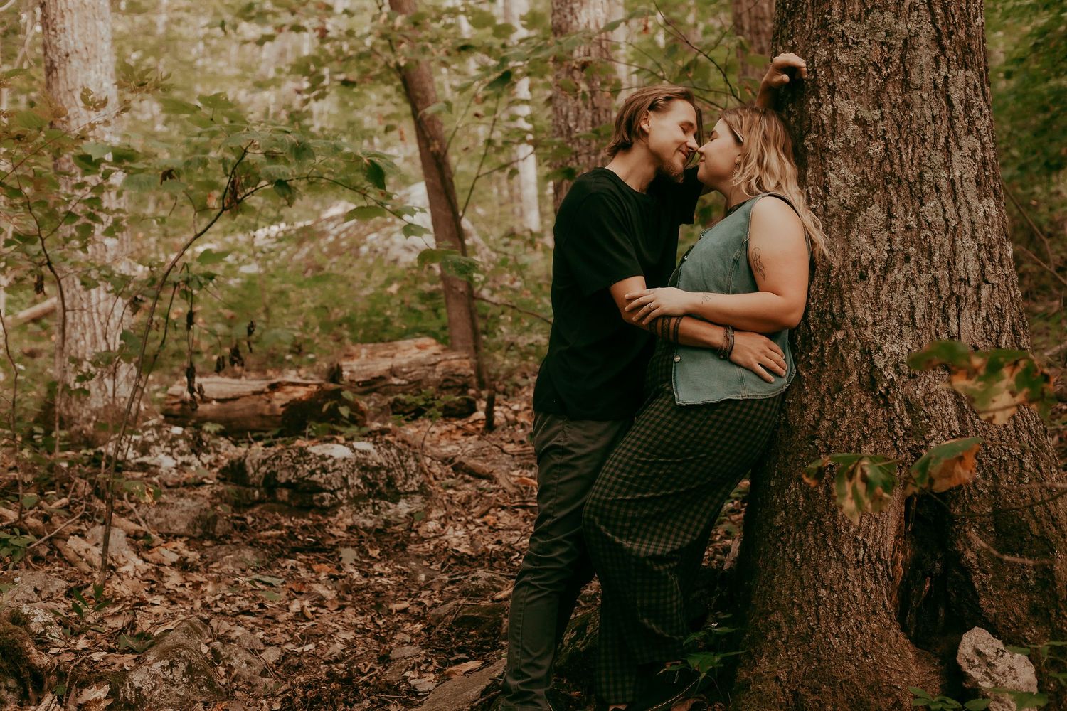 Romantic couple kissing while leaning against large tree trunk in woodland forest.