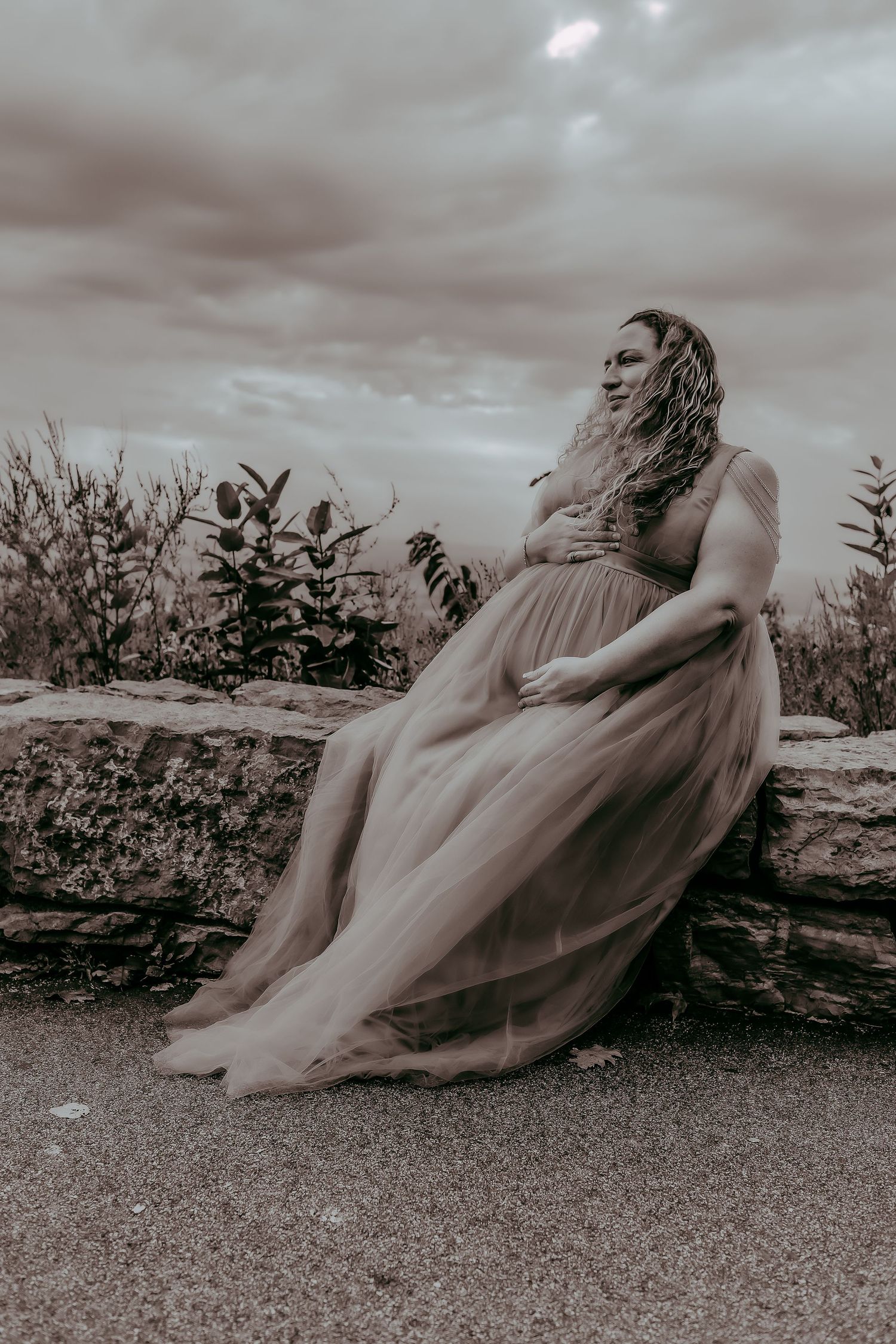 Black and white photo of pregnant woman in tulle gown sitting on stone wall outdoors.