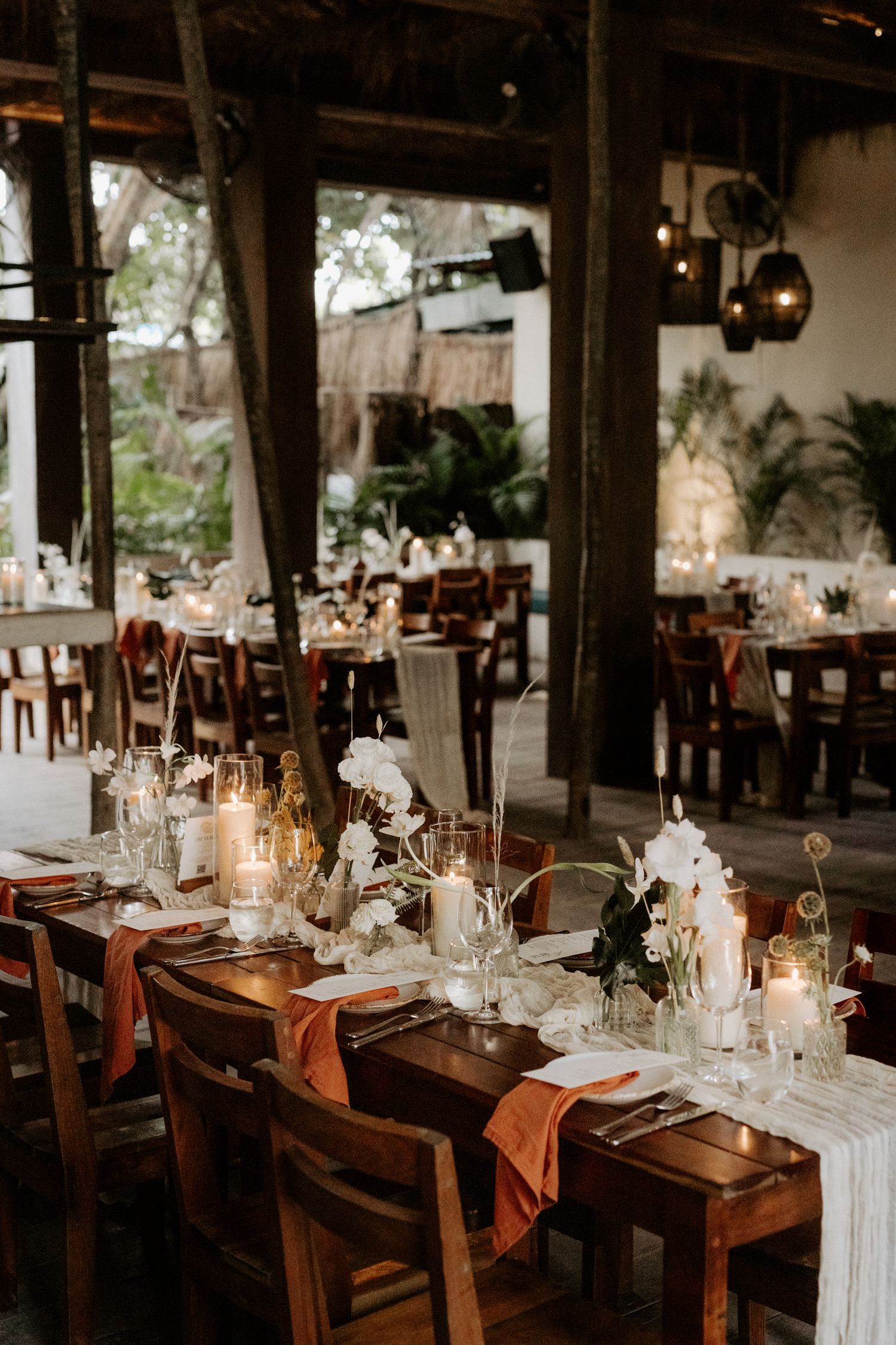 Boho-elegant Tulum reception venue with wooden tables, hanging greenery and ambient lighting creating intimate atmosphere for luxury wedding