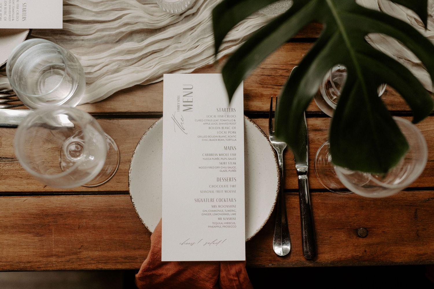 Elegant Tulum destination wedding table setting with minimalist menu card, white plate, crystal glassware and tropical leaf accent on wood