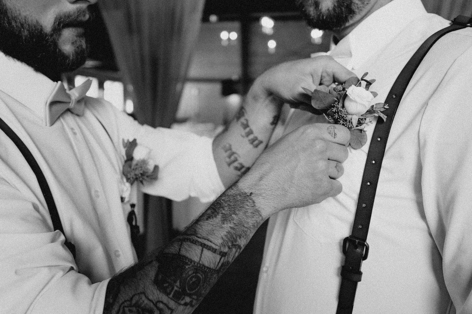 Black and white close-up of best man adjusting groom's boutonnière, cinematic documentary approach capturing pre-ceremony details
