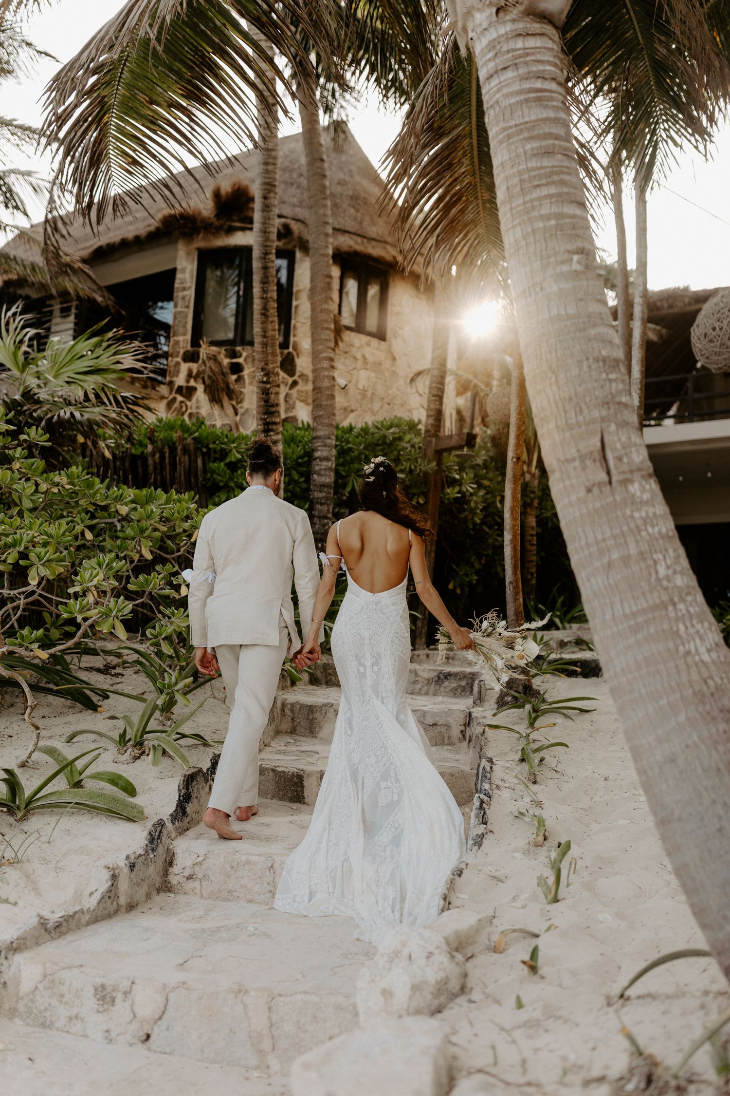 Bride and groom walking hand in hand on sandy path toward luxury beachfront villa with palm trees and sunlight in Tulum destination wedding