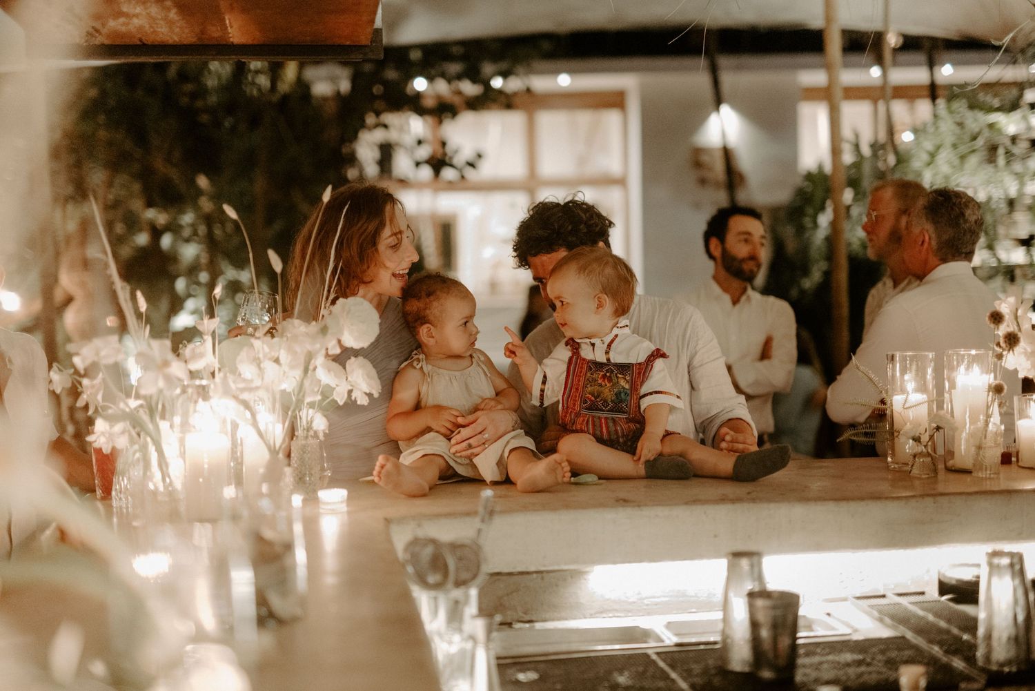 Intimate Tulum wedding reception with kids sharing moment at candlelit table, surrounded by greenery and warm lights in luxury Mexico venue