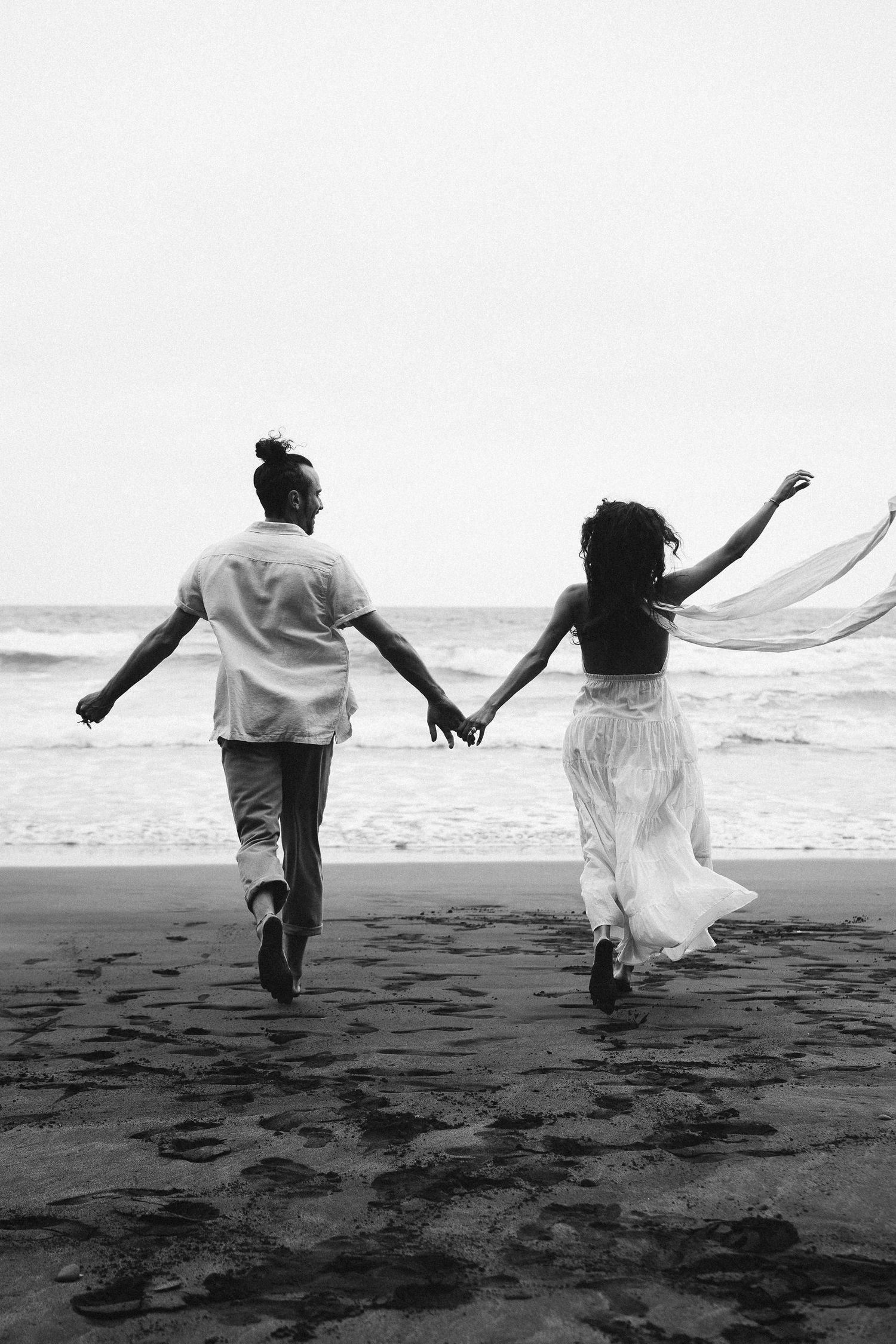 Black and white 35mm film beach portrait of couple running hand-in-hand in Tenerife, Spain, cinematic destination engagement photography
