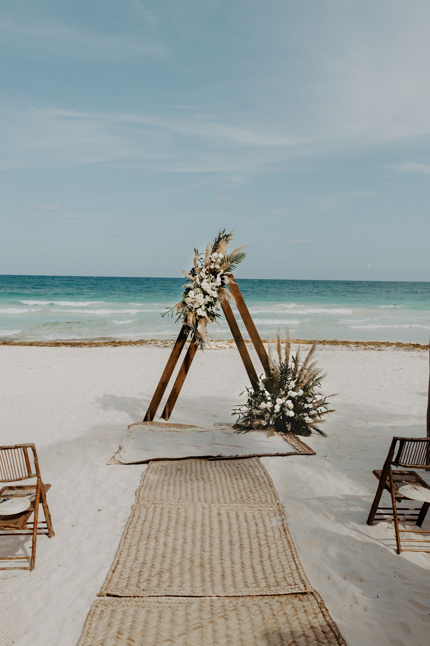 Elegant beachfront wedding ceremony arch with natural florals on sand beach in Tulum, featuring minimalist boho luxury destination design