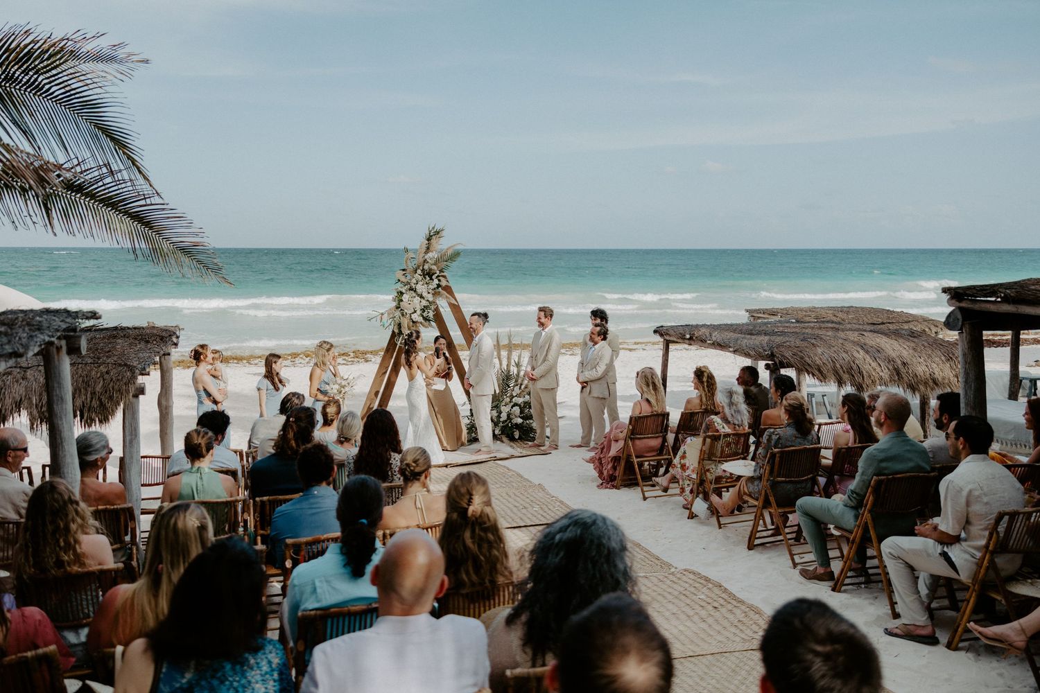 Tulum beachfront wedding ceremony with wooden triangle arch, guests seated on both sides of aisle, turquoise ocean backdrop and palm trees