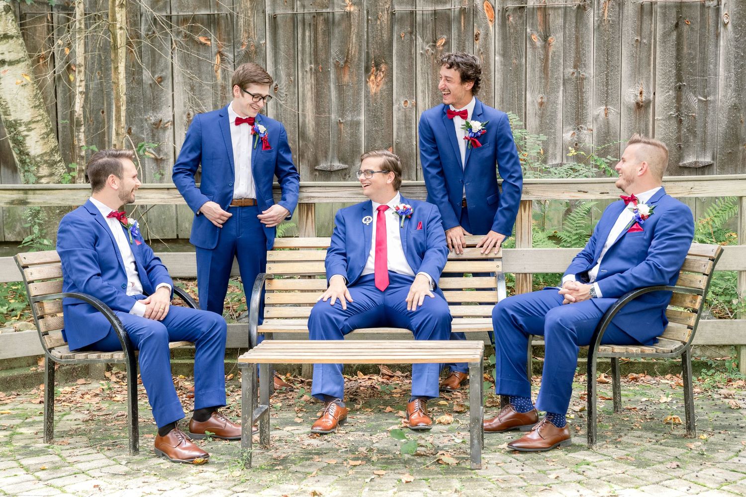 Groomsmen in matching navy blue suits and brown shoes sit and stand casually on wooden benches for wedding photos.