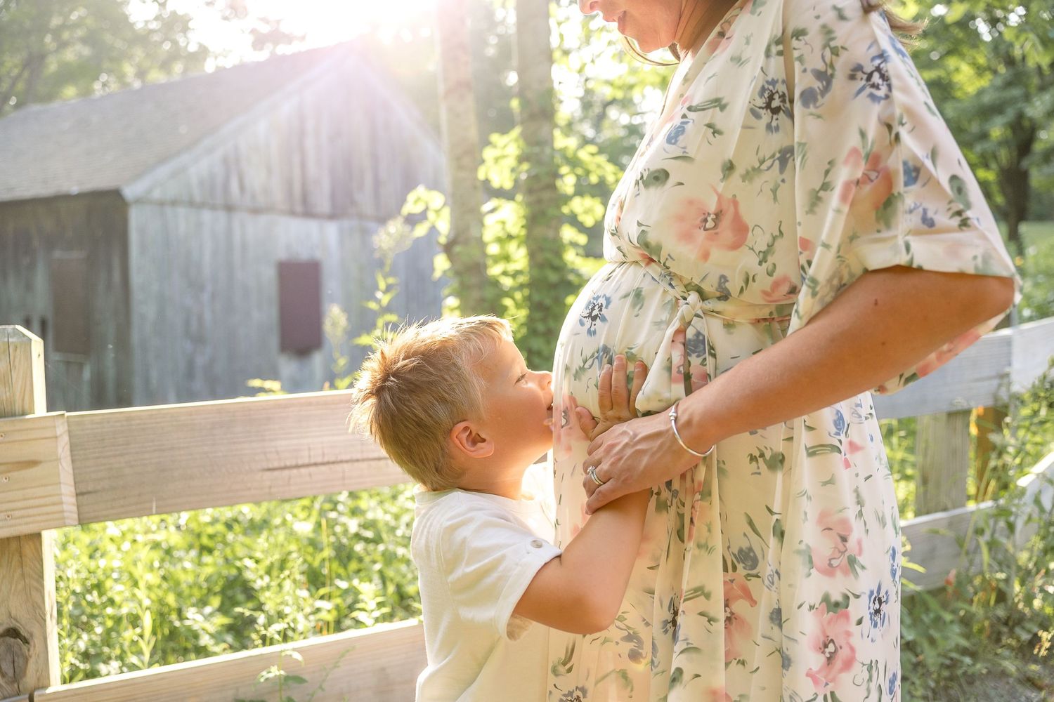 A young child touches a pregnant woman's belly while standing on a wooden fence in glowing sunlight.