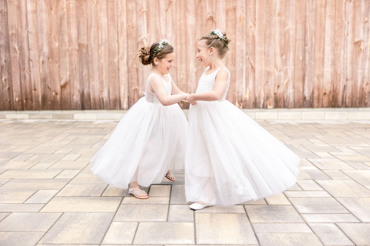 Two flower girls in white dresses twirling together on stone patio.