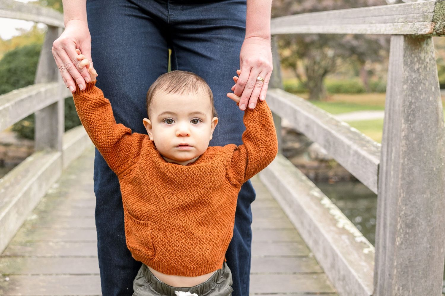 Small child in orange sweater holds hands while standing on wooden bridge with caregiver.