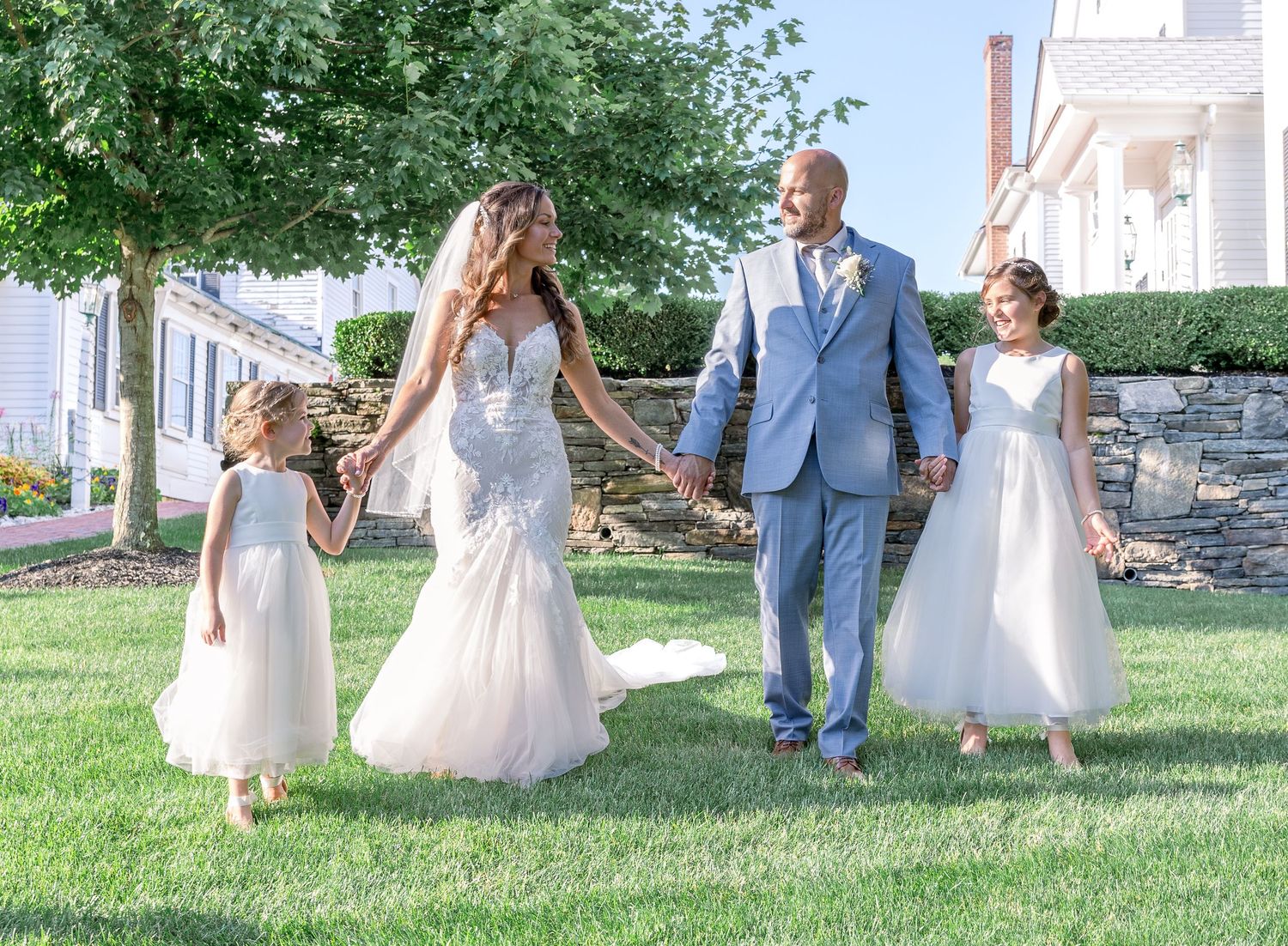 A wedding party walks together on a green lawn holding hands in elegant formal attire on a sunny day.
