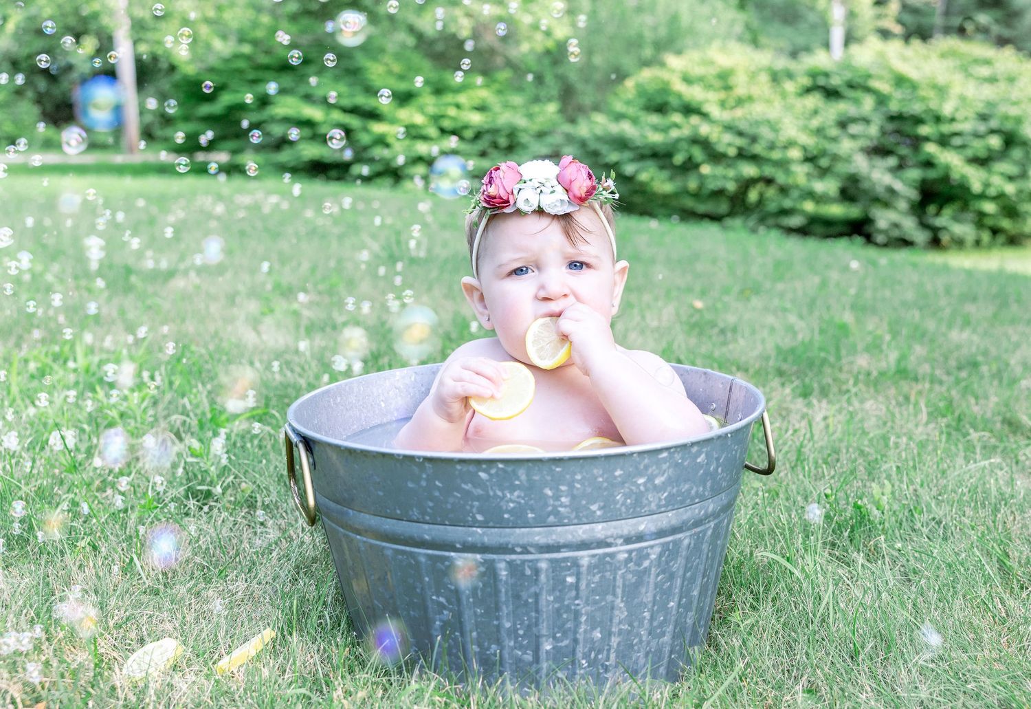 A baby wearing a pink bow sits in a galvanized metal tub outdoors surrounded by dandelion seeds on a summer day.