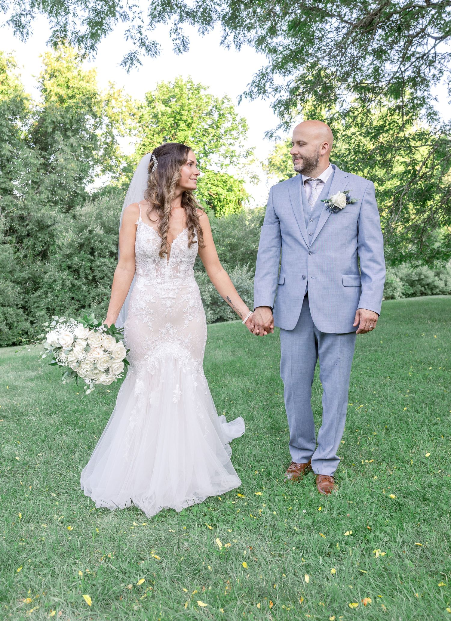 Newlywed couple holding hands outdoors in a garden setting, bride in lace gown and groom in light gray suit.