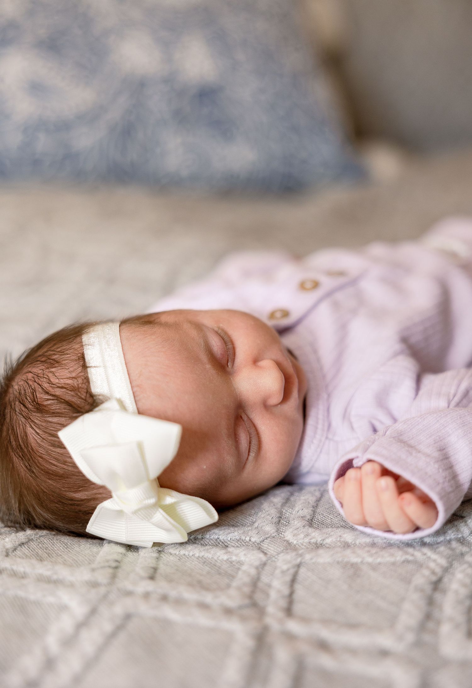 Sleeping newborn baby wearing pink outfit and white bow headband on gray blanket.
