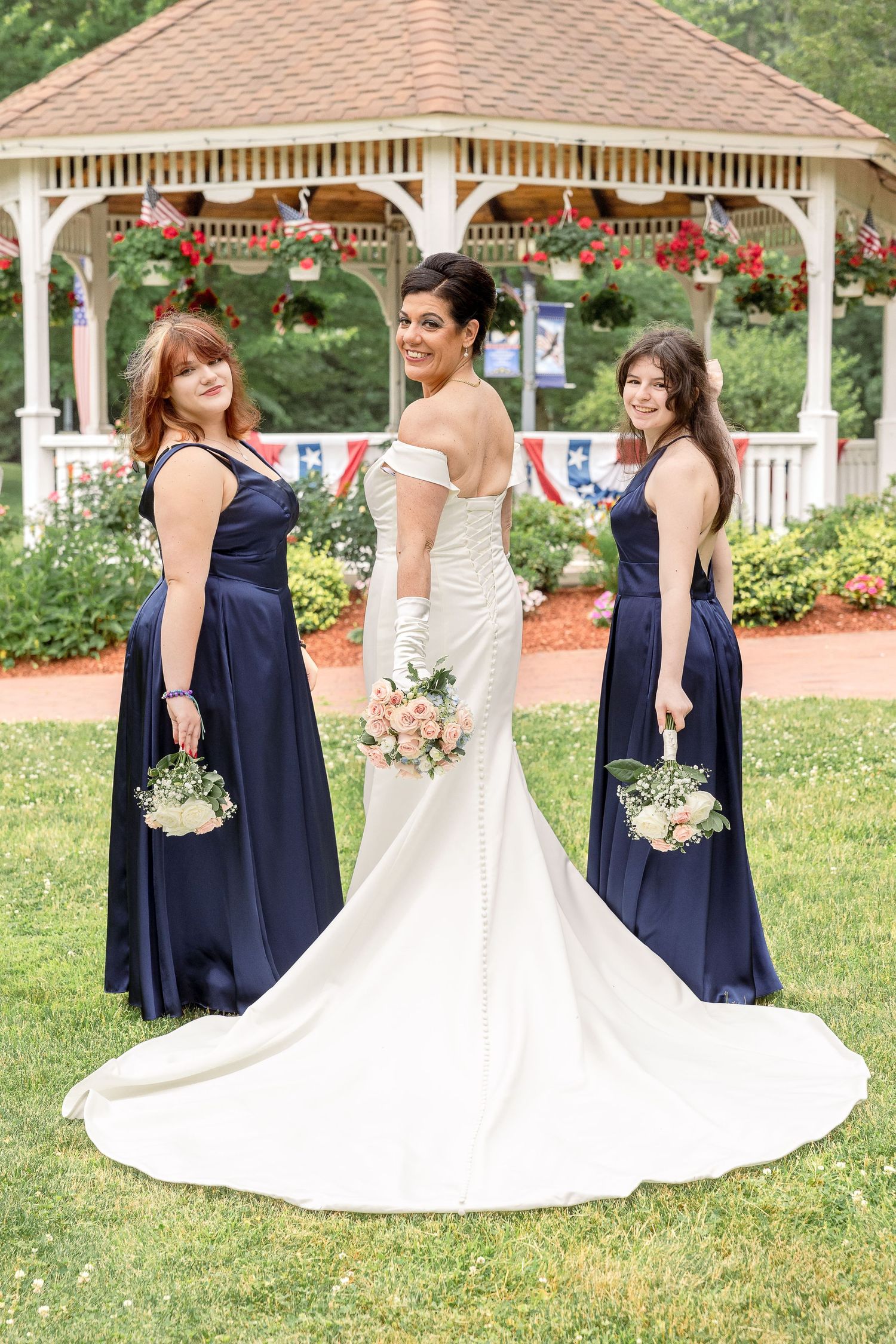 Wedding party posing in front of garden gazebo with bride in white gown and bridesmaids in navy blue dresses.