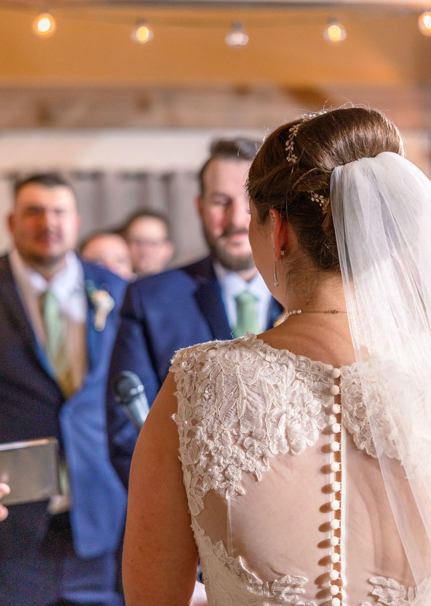 A bride in a lace wedding dress with button details and veil stands at the altar during a wedding ceremony.
