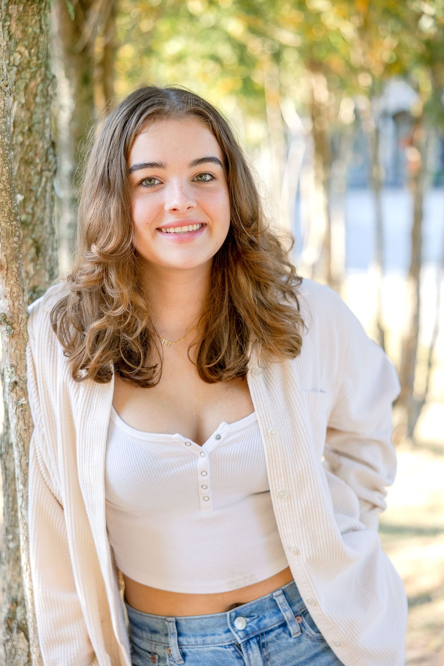 A young woman in a cardigan and jeans stands among trees on a sunny day.