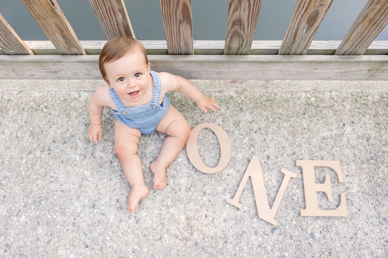 Baby in blue outfit lying on concrete floor next to wooden ONE letters.
