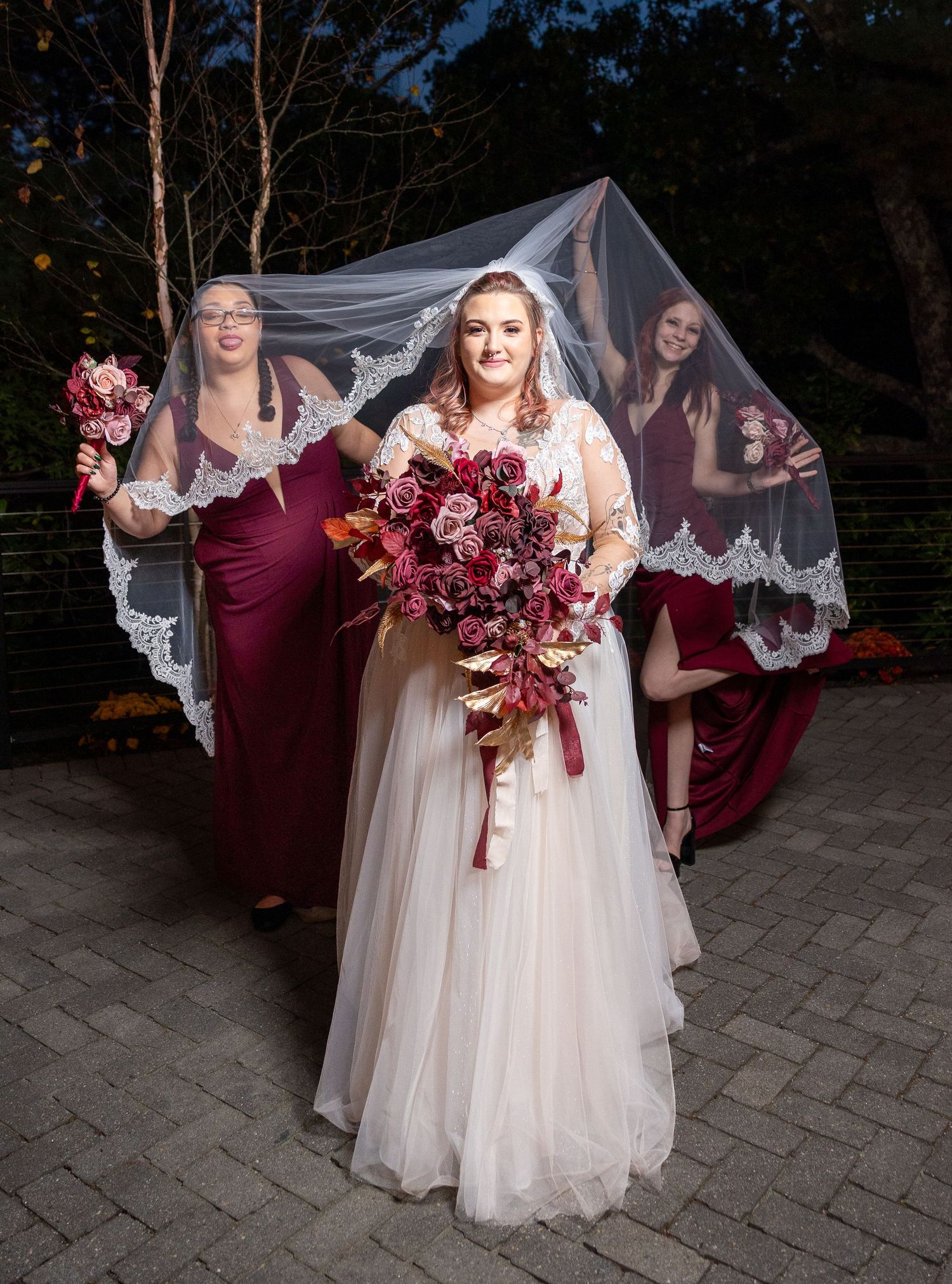 A wedding party in burgundy and white dresses poses outdoors as a lace veil blows dramatically in the wind.
