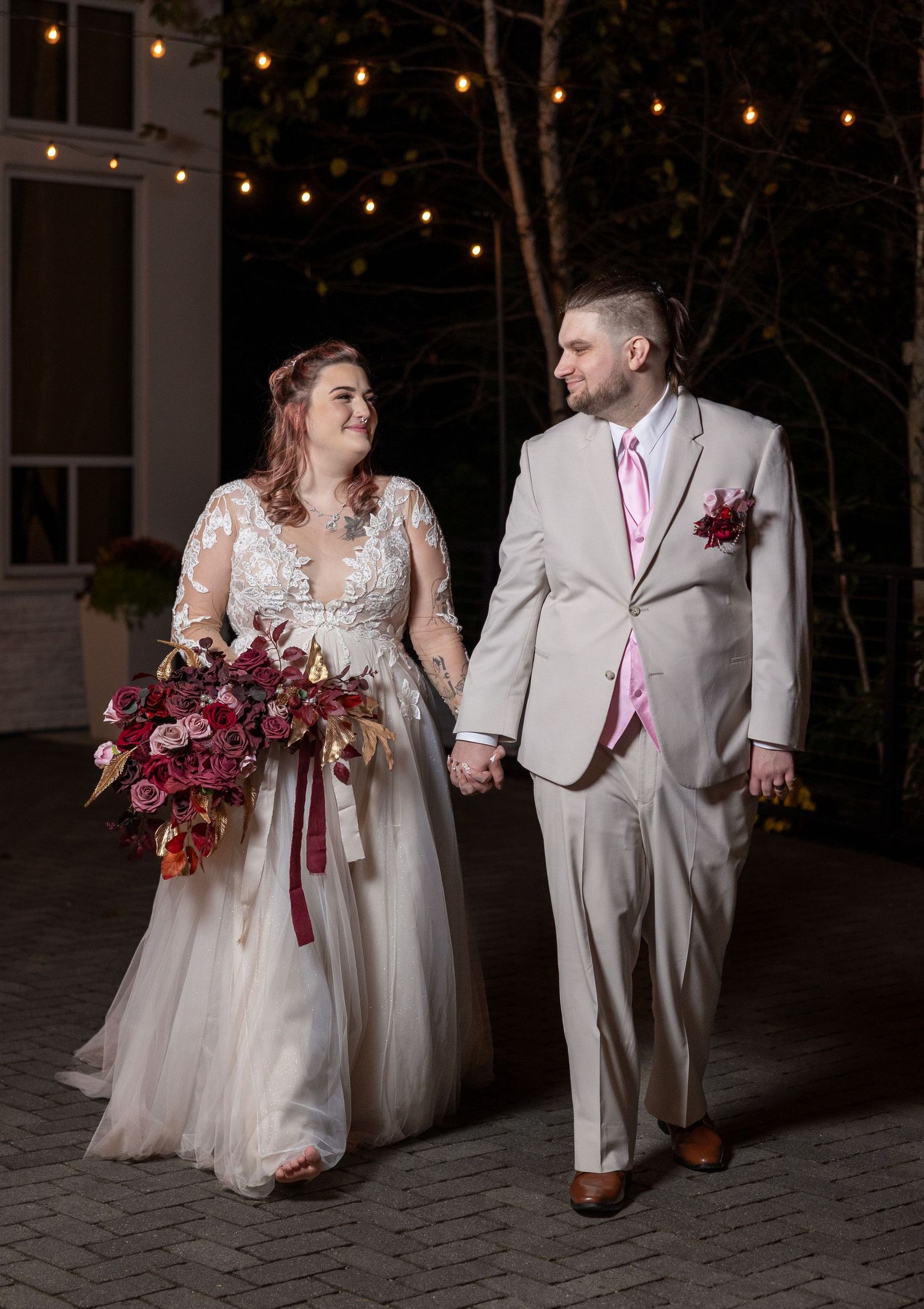 Newlywed couple walk together at night under string lights with bride holding burgundy bouquet and wearing lace gown.