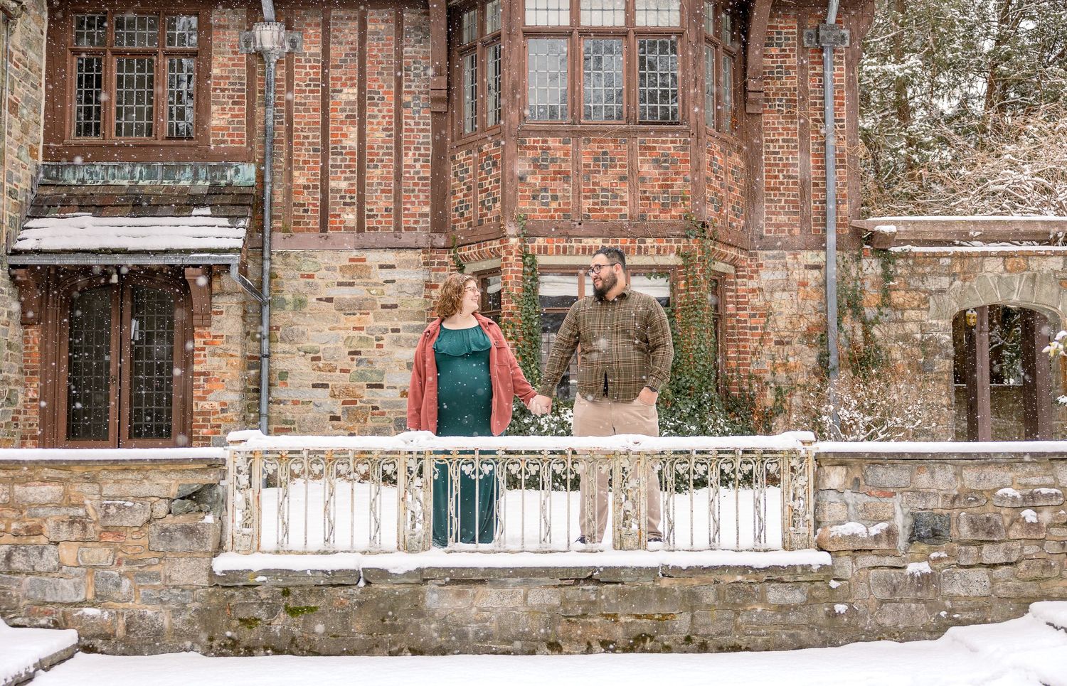 A couple stands on a snow-covered stone balcony in front of a rustic brick building during winter.