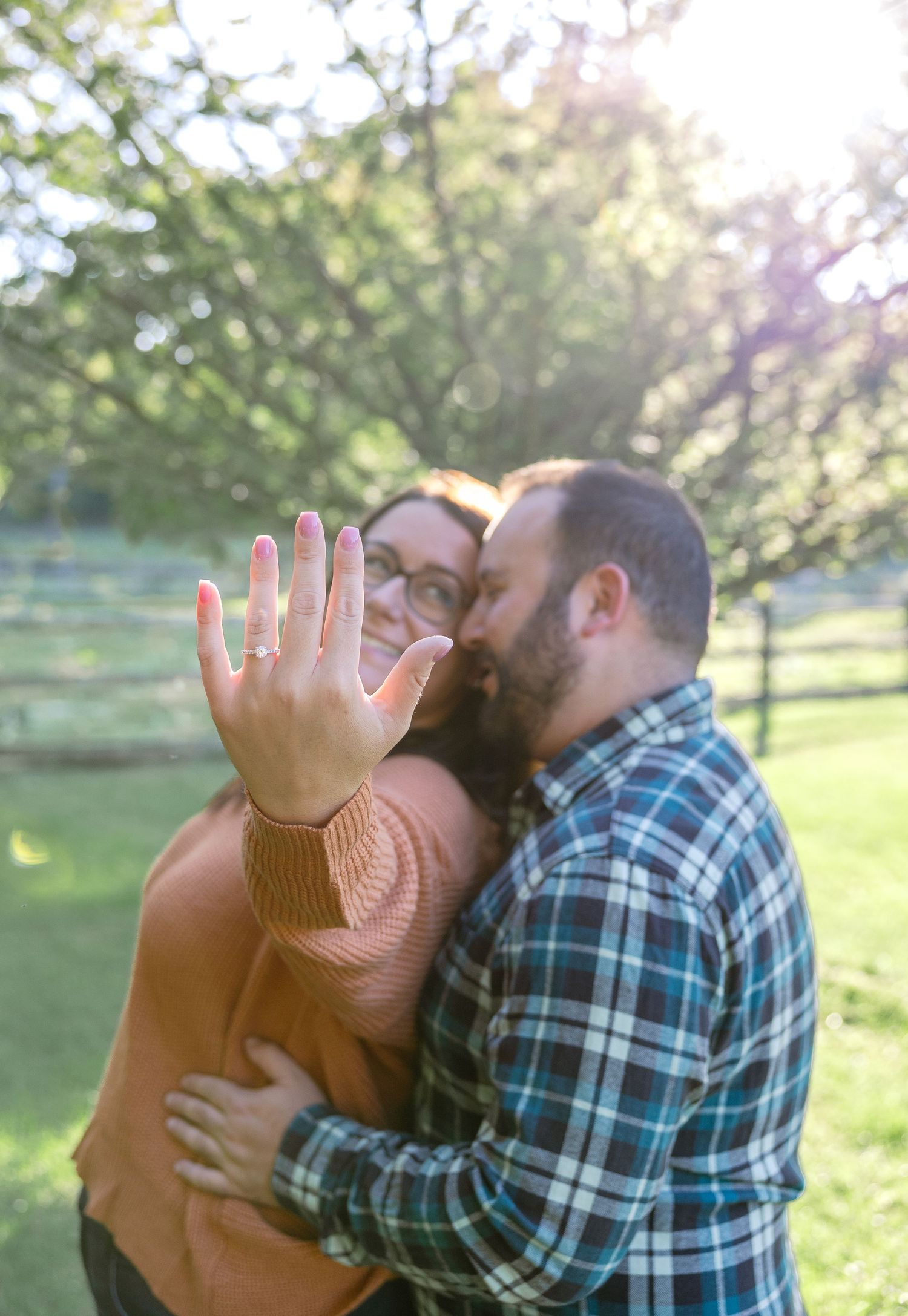 A couple embraces outdoors in a sunny park setting as they show off a ring in a joyful engagement moment.