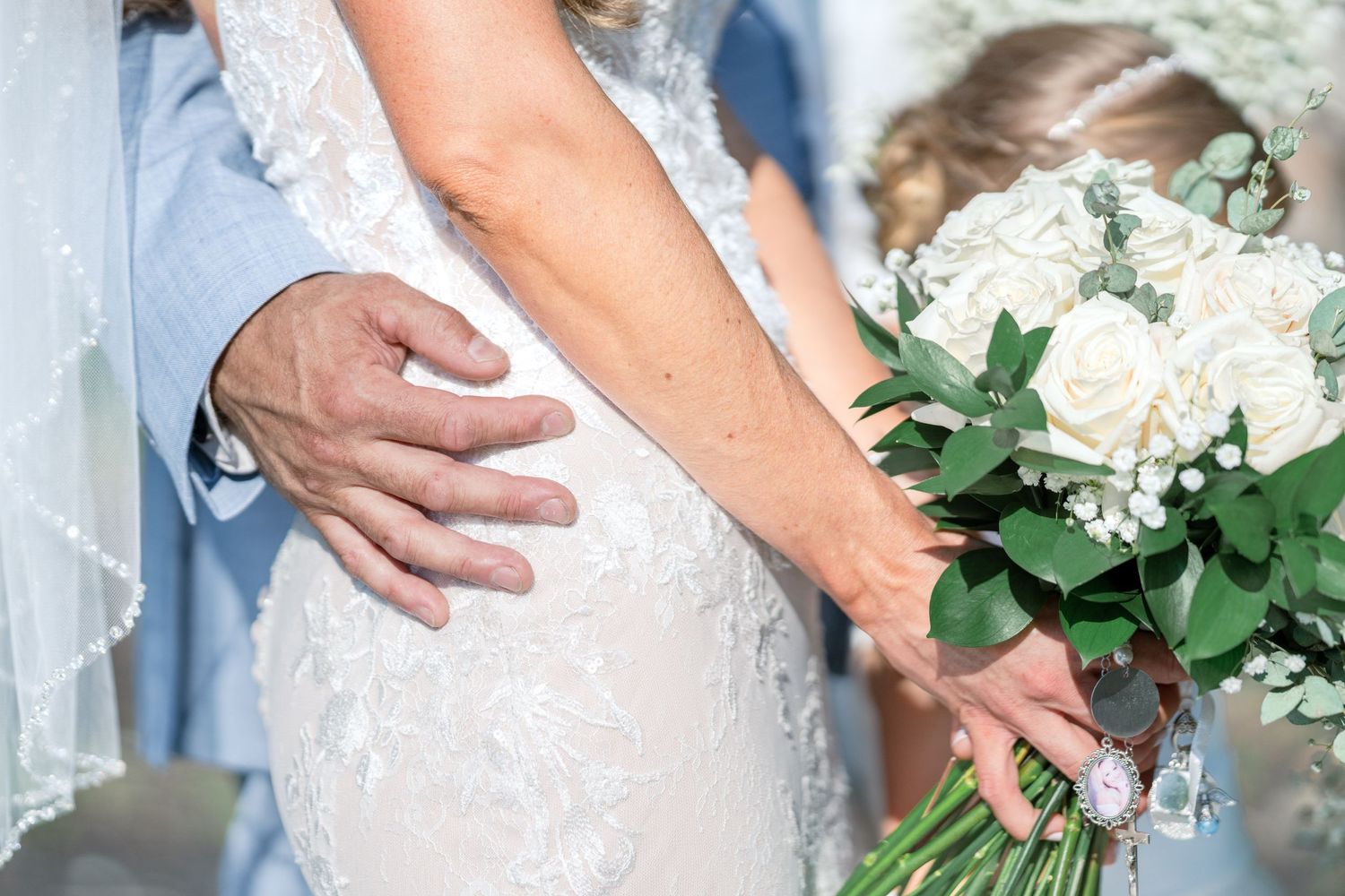 A close-up view of a white lace wedding dress with hands holding a bouquet of white roses and greenery.