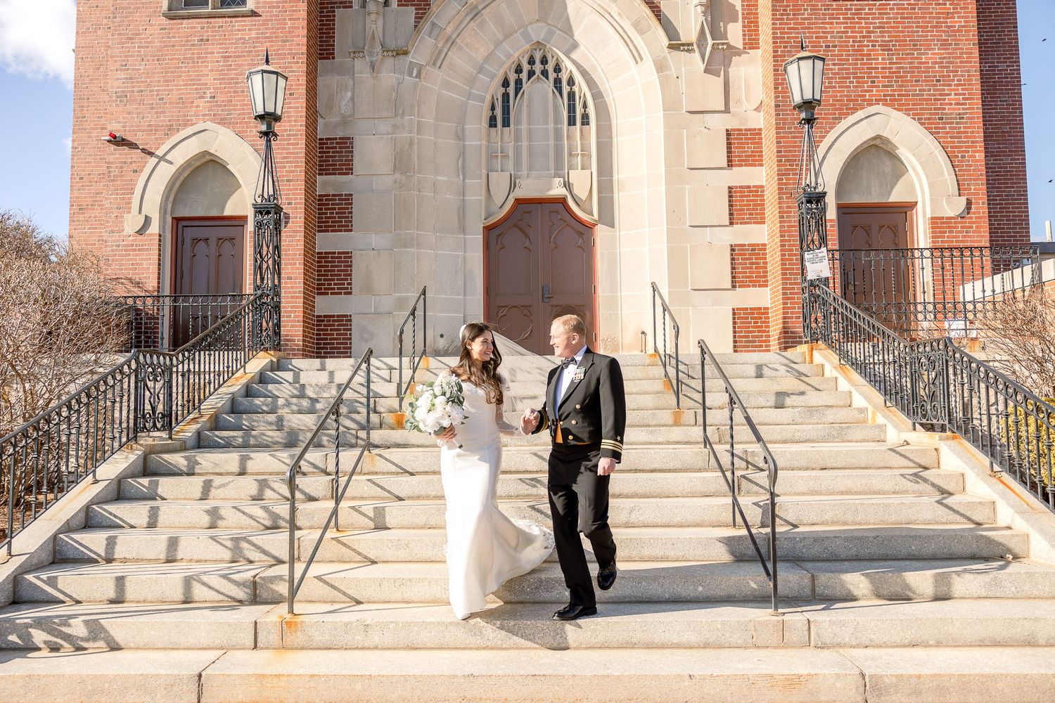 A wedding couple descends the grand stone steps of a red brick church with Gothic arched doorways and decorative railings.