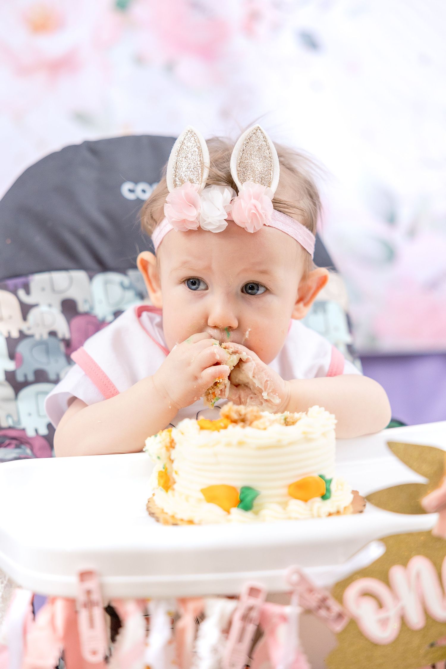 A baby wearing bunny ears headband tastes birthday cake from a white plate with pink party decorations in background.