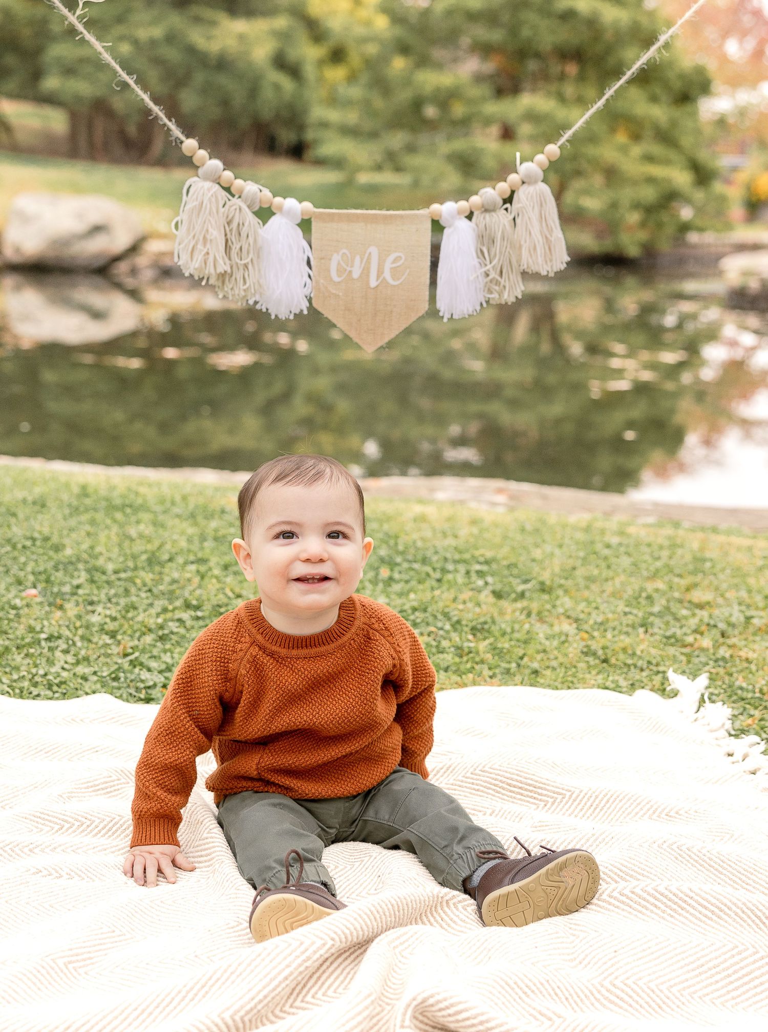Baby in a rust-colored sweater and gray pants sits on a blanket outdoors with a 'One' banner hanging above.