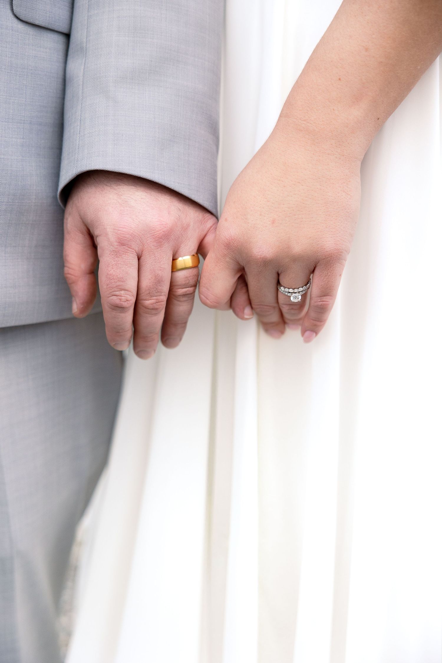 Close-up of newlywed couple's hands showing wedding rings against white dress and gray suit.