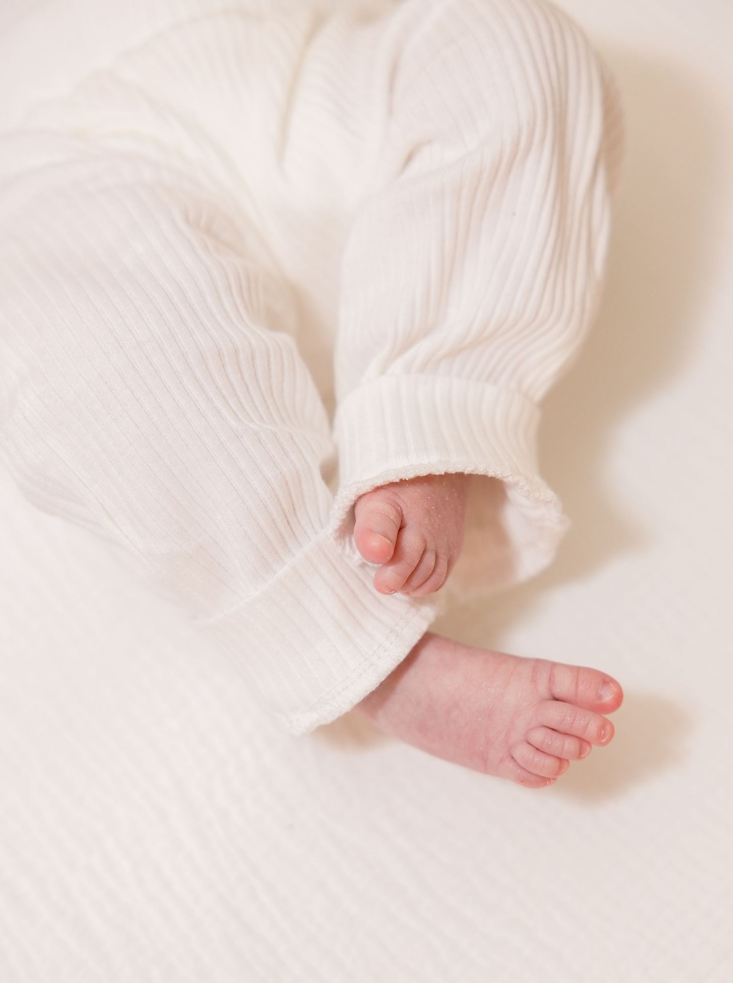 Tiny bare baby feet and legs peek out from soft white knitted fabric in a minimalist nursery photo.