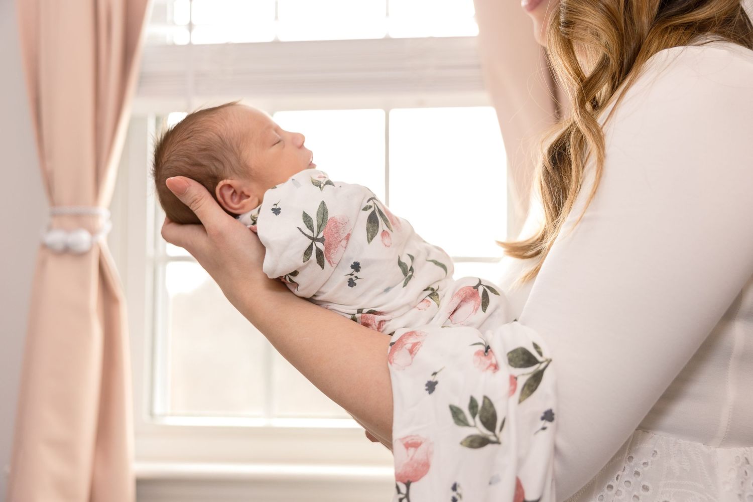 A baby wrapped in a floral print swaddle blanket is held up near a sunlit window with pink curtains in the background.